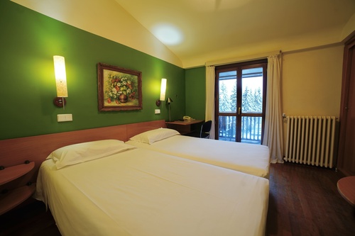 Acogedora habitación de hotel con dos camas individuales, escritorio y balcón con vista a árboles nevados.