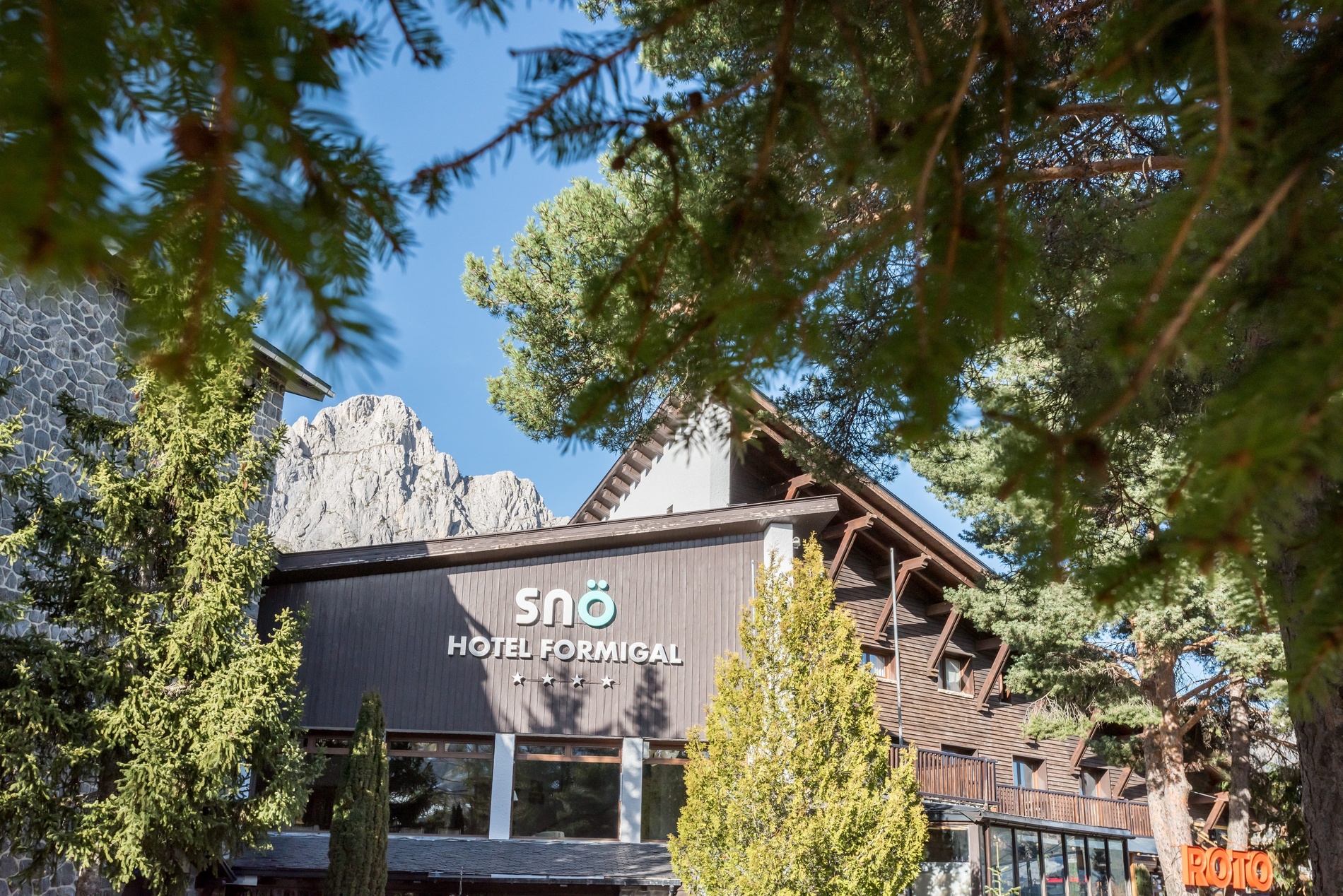 Hotel Snö Formigal en un entorno de montaña con arquitectura de madera, árboles y cielo azul.