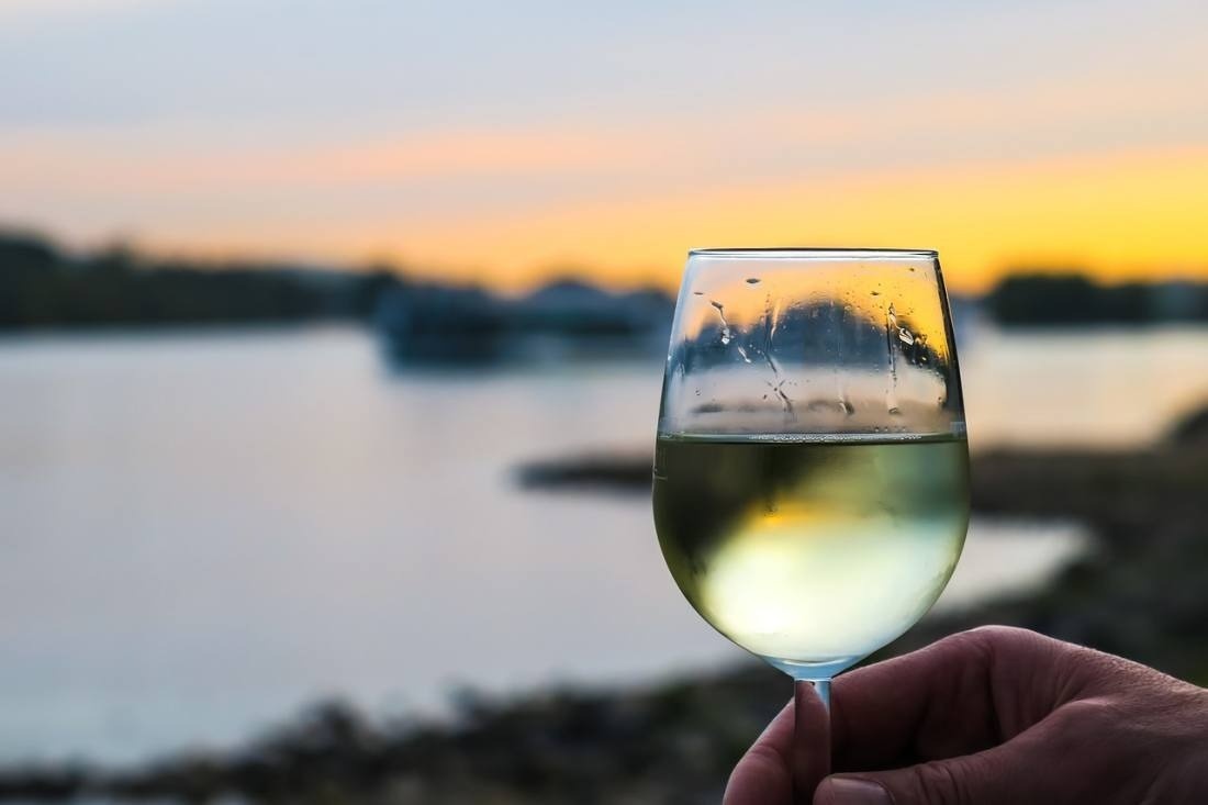 una persona sostiene una copa de vino frente a un río al atardecer