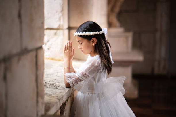 una niña con un vestido blanco y una corona de flores está rezando en una ventana .