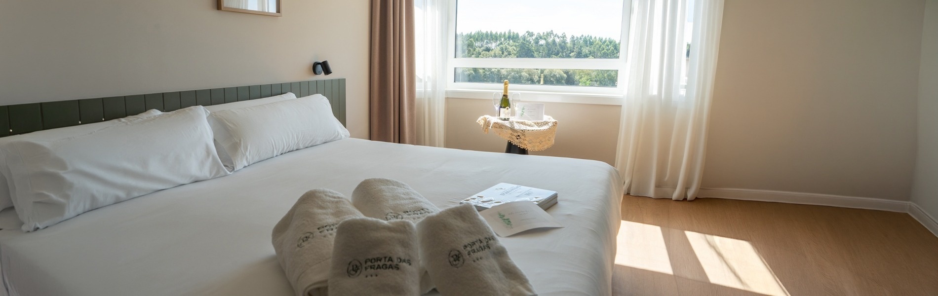 Habitación de hotel luminosa con cama, zapatillas, champagne y vistas verdes.