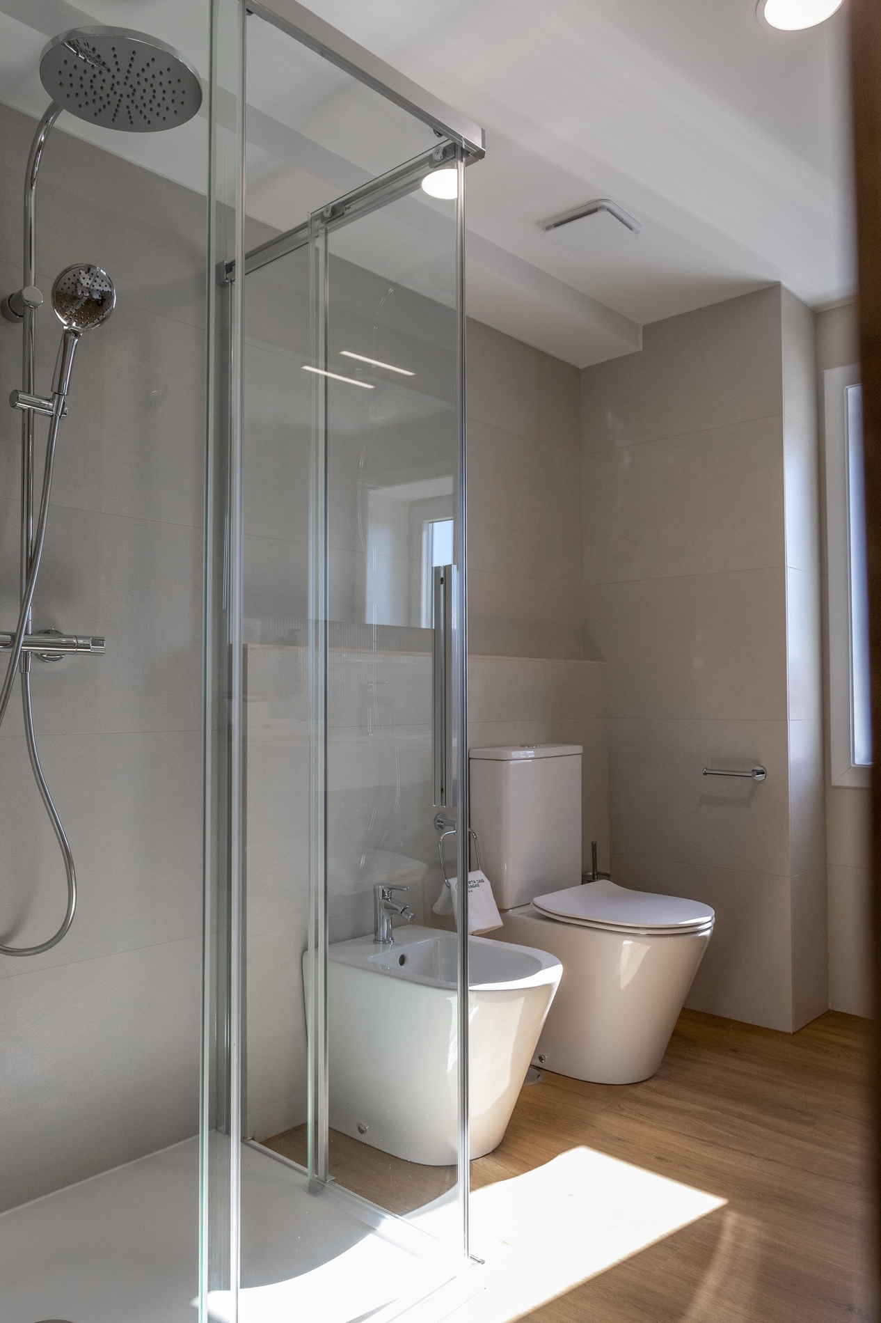 Une salle de bain moderne et lumineuse, comprenant une cabine de douche en verre, un bidet et des toilettes, avec des murs carrelés clairs et un sol en bois.