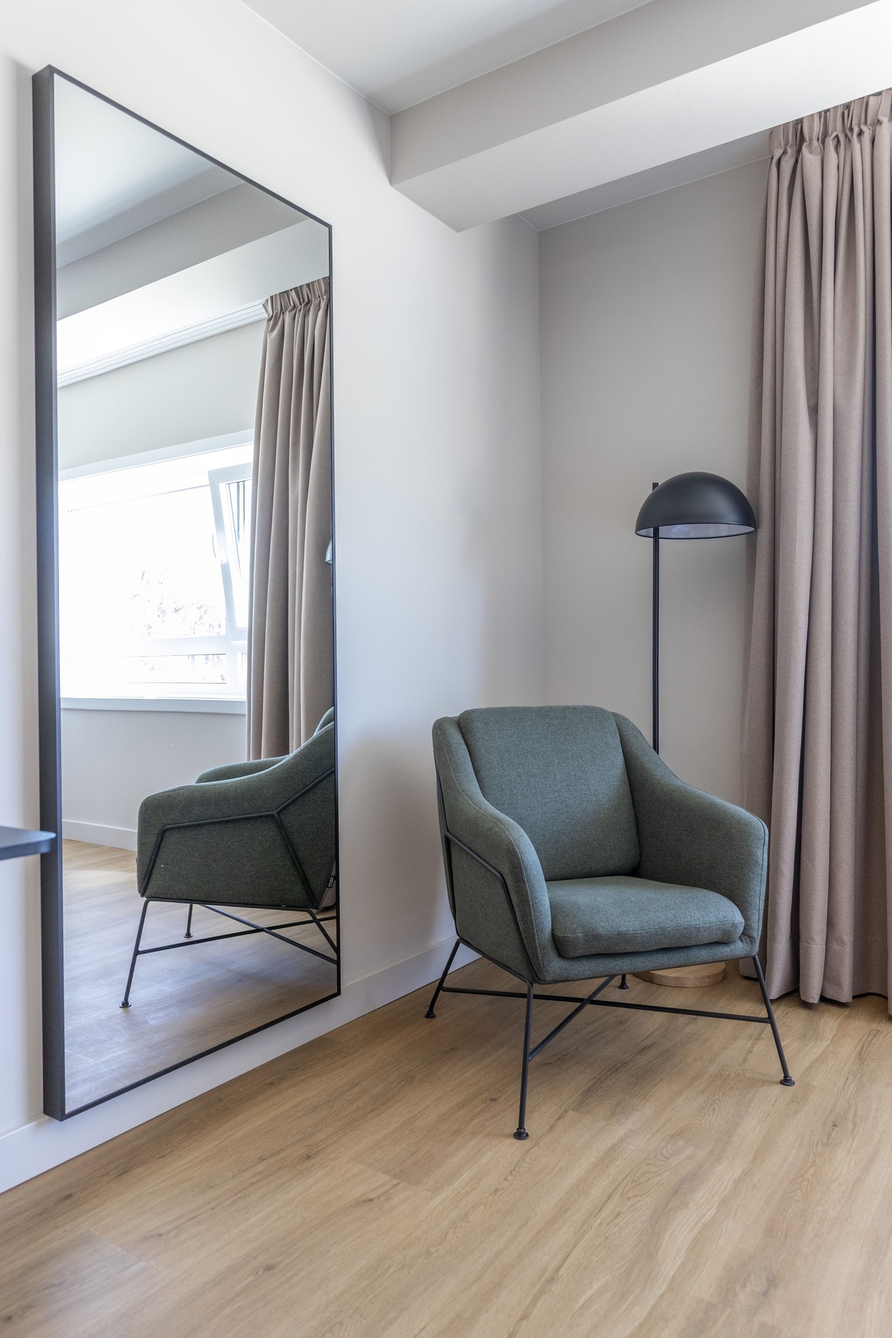Un fauteuil vert moderne est placé dans un coin de pièce avec un grand miroir mural reflétant une fenêtre, des rideaux beiges et un lampadaire noir sur un parquet en bois.