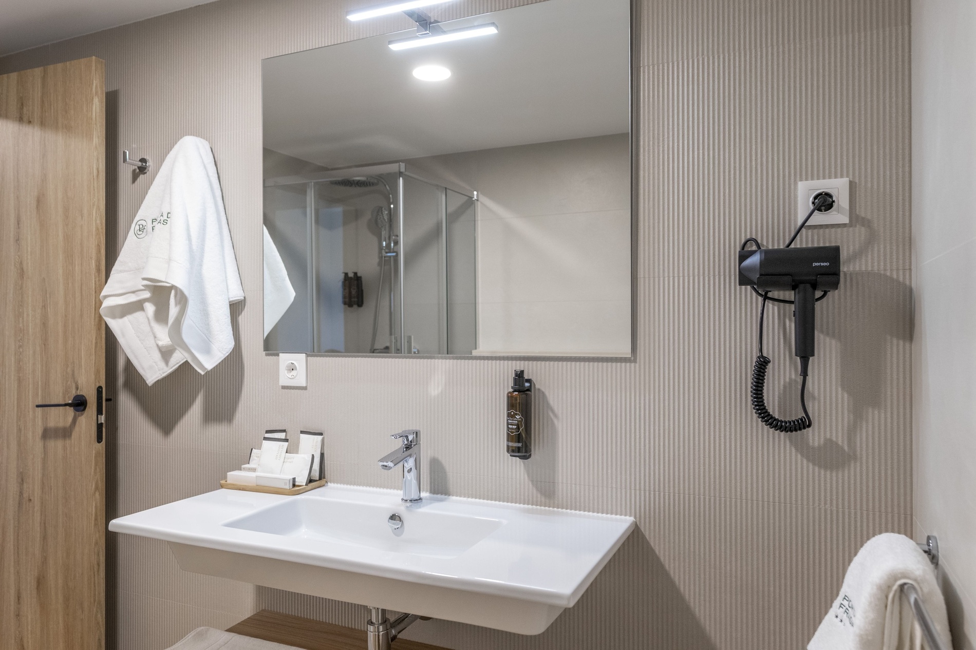 Une salle de bain moderne est présentée avec un lavabo, un grand miroir reflétant une cabine de douche, un sèche-cheveux mural et des articles de toilette, le tout sur des murs carrelés à motifs striés.