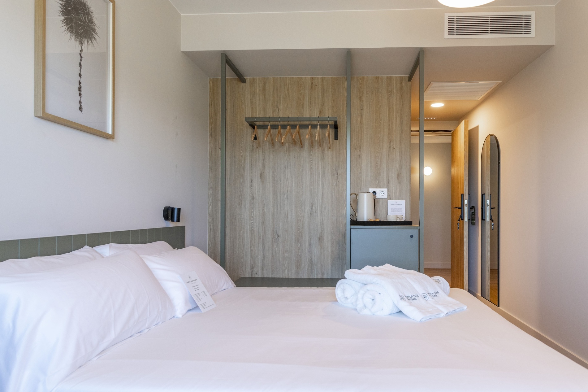 Une chambre d'hôtel moderne est présentée avec un lit fait de draps blancs, une petite penderie ouverte, une bouilloire sur une petite console et un miroir mural incurvé.