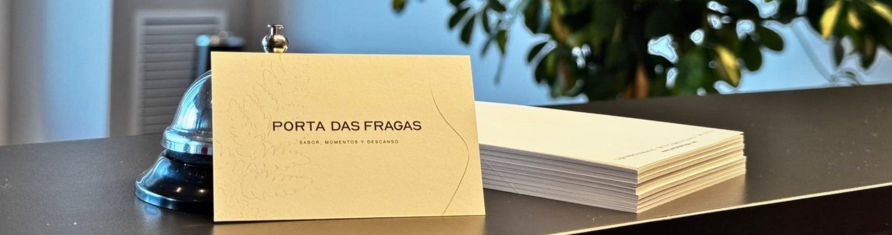 Sobre un mostrador oscuro, una tarjeta con el texto "PORTA DAS FRAGAS" y una campanilla de recepción se ven frente a una planta verde borrosa.
