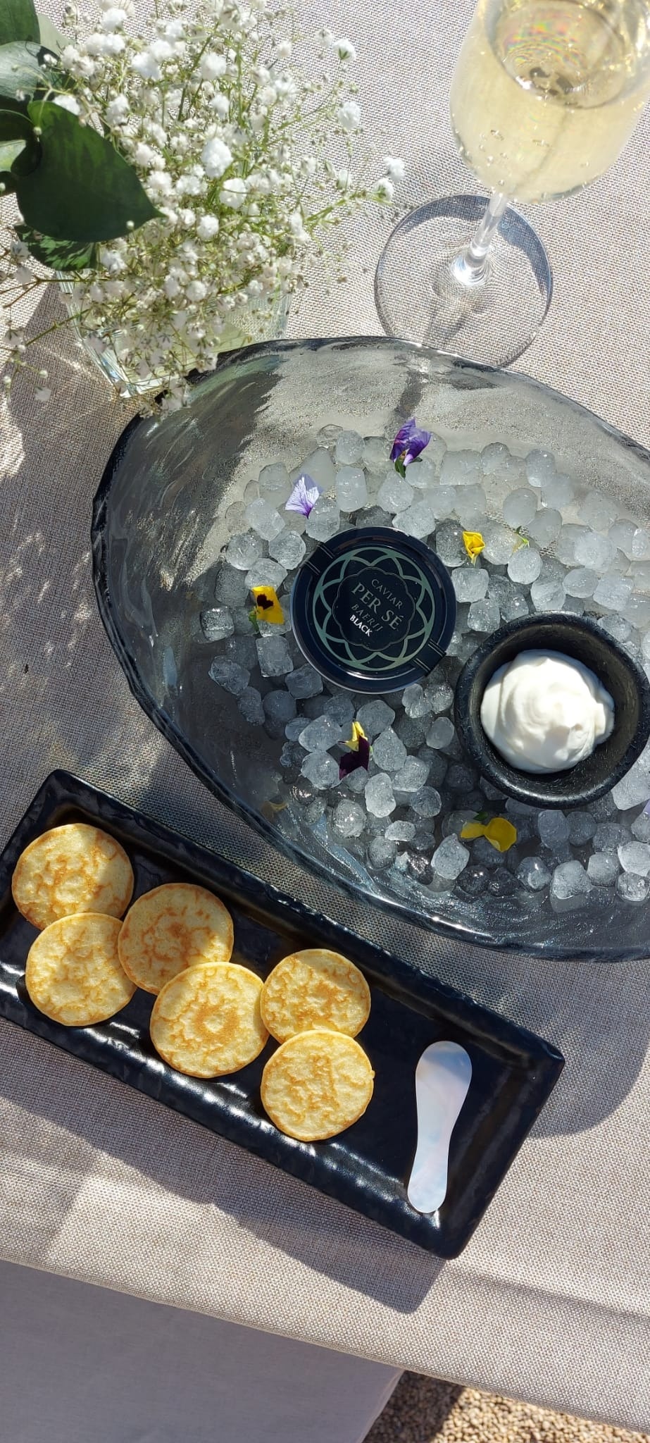 Kaviar auf Eis, Blinis, Crème fraîche und ein Sektglas sind elegant mit weißen Blumen auf einem Tisch angerichtet.