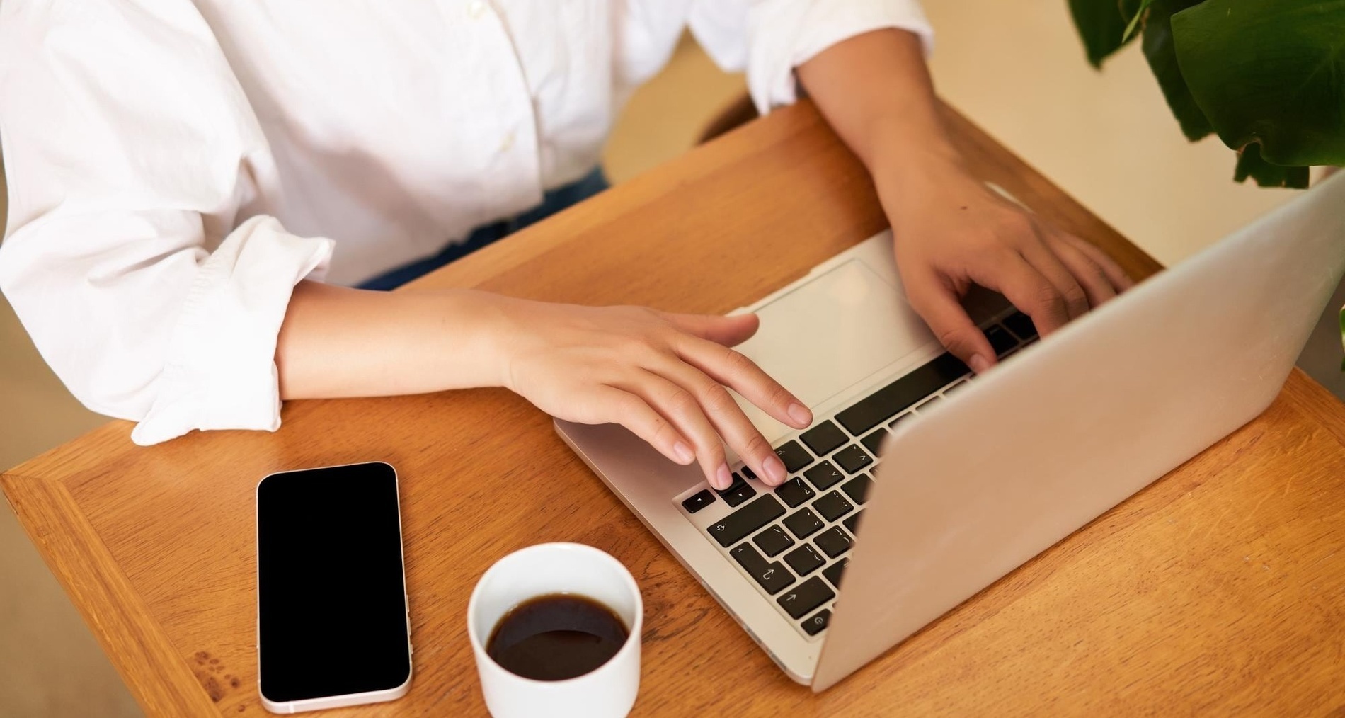 una persona está escribiendo en una computadora portátil junto a un teléfono y una taza de café