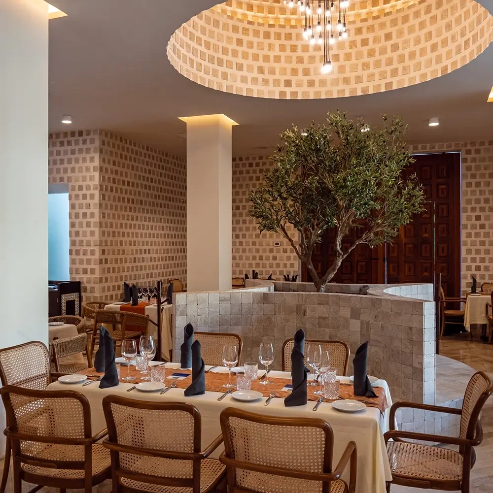 Elegant dining room with rattan chairs, a beautifully set table, and a live tree, ideal for your Sunset World Resorts: Corporativa experience.