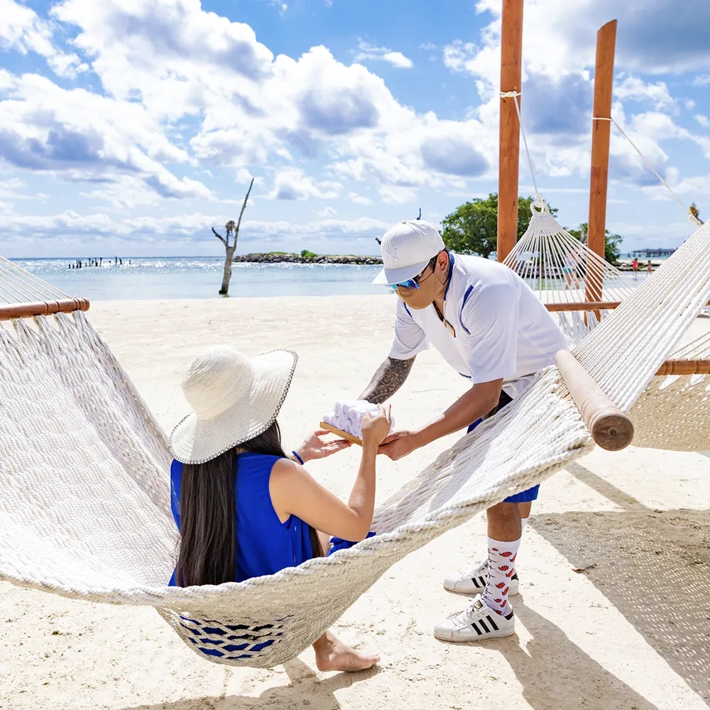 A server offers Sunset Hotels Collection: Corporativa to a guest relaxing in a hammock on a beautiful beach.