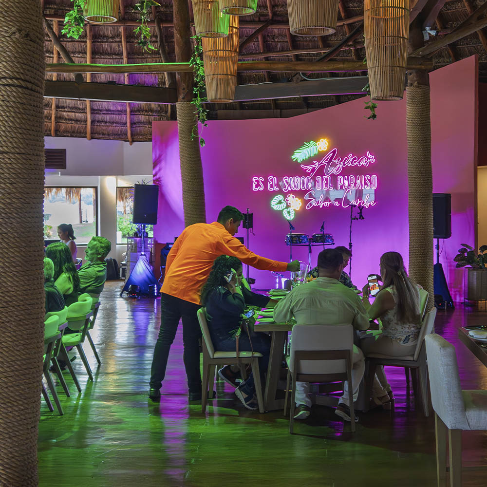 Restaurant interior with neon sign Sunset Hotels Collection: Corporativa, people dining, and tropical decor.