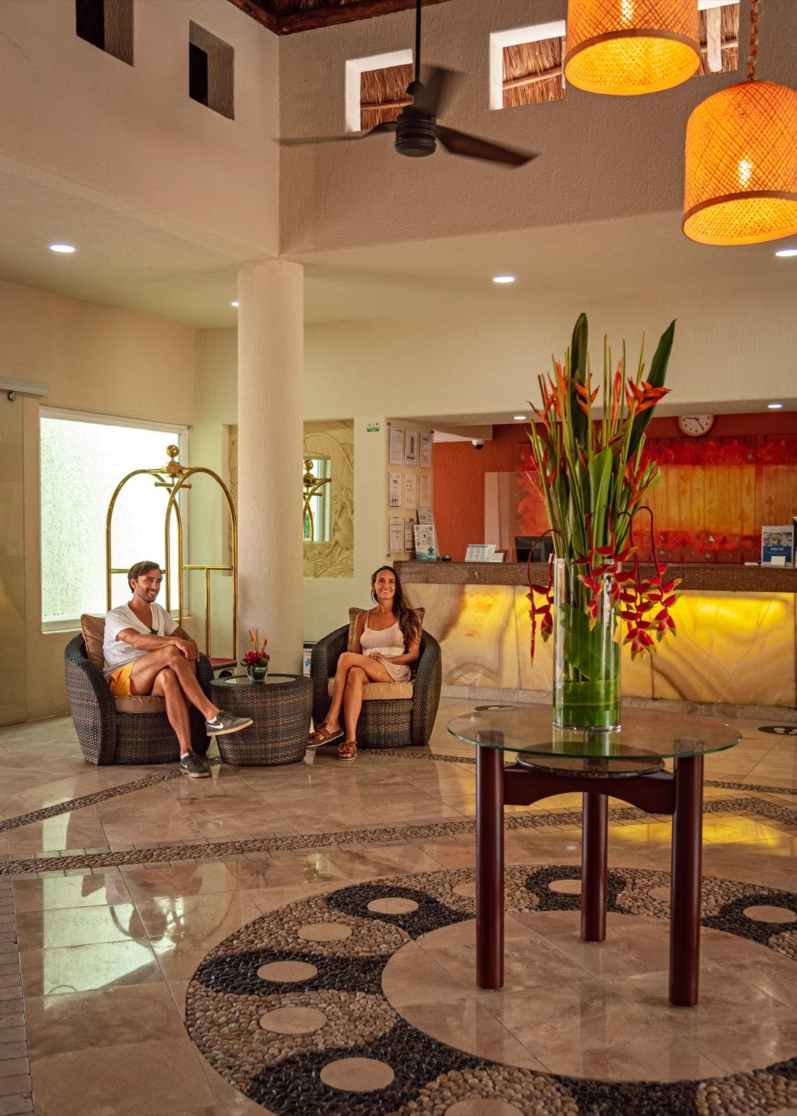 Couple relaxing in hotel lobby lounge area with tropical decor and front desk in background Laguna Suites | Boutique Hotel in Cancun | Official Site.