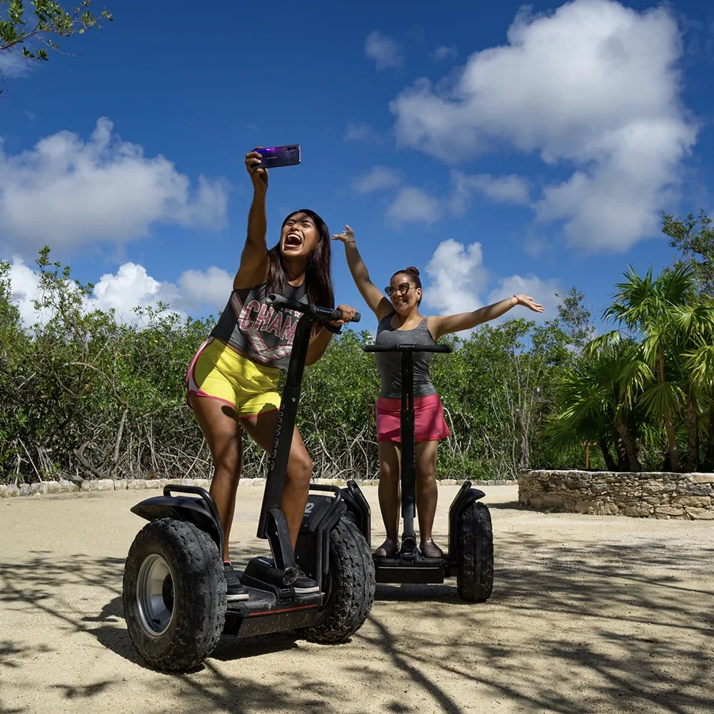 Two women on Segways Sunset Hotels Collection: Corporativa take a selfie in a tropical resort with palm trees and blue skies.