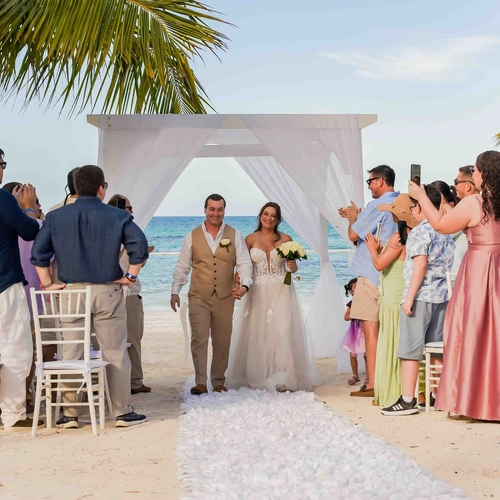 Novios caminando por el pasillo de una ceremonia en la playa rodeados de invitados