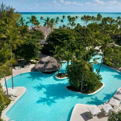 Vista aérea de la piscina con isla de palmeras y el mar al fondo en Impressive Punta Cana