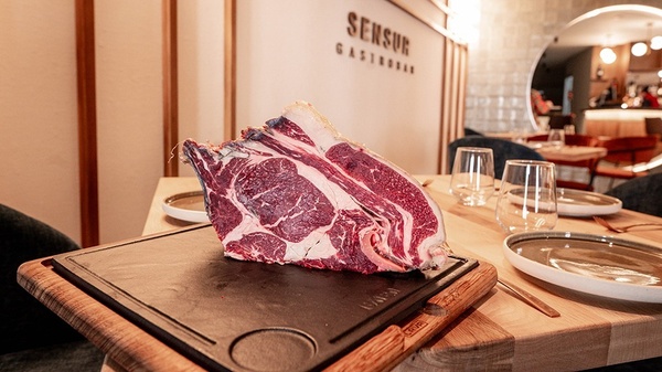 a large piece of meat sits on a cutting board in front of a wall that says sensur gastrobar