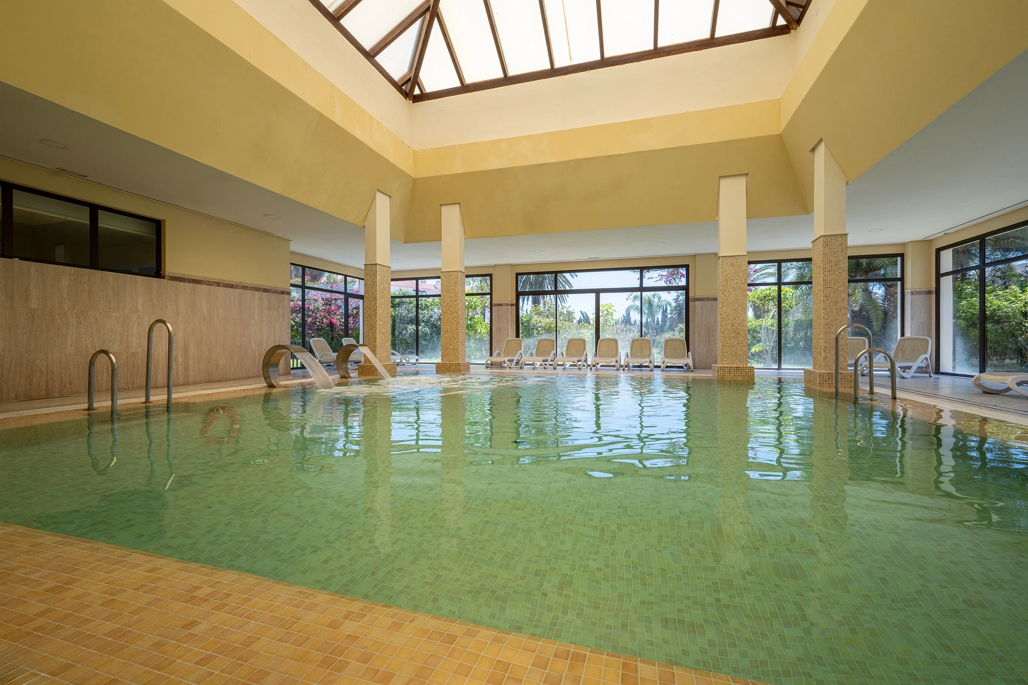 Piscina climatizada cubierta del spa con claraboya, tumbonas y ventanales al jardín