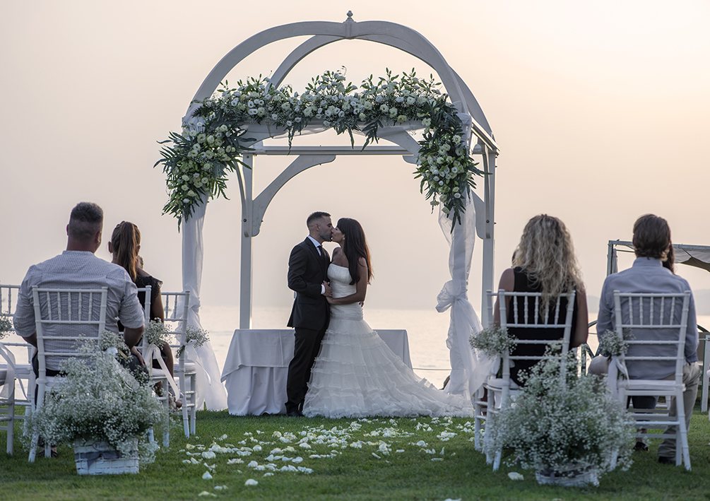 Novios besándose bajo un arco de flores en una ceremonia al atardecer