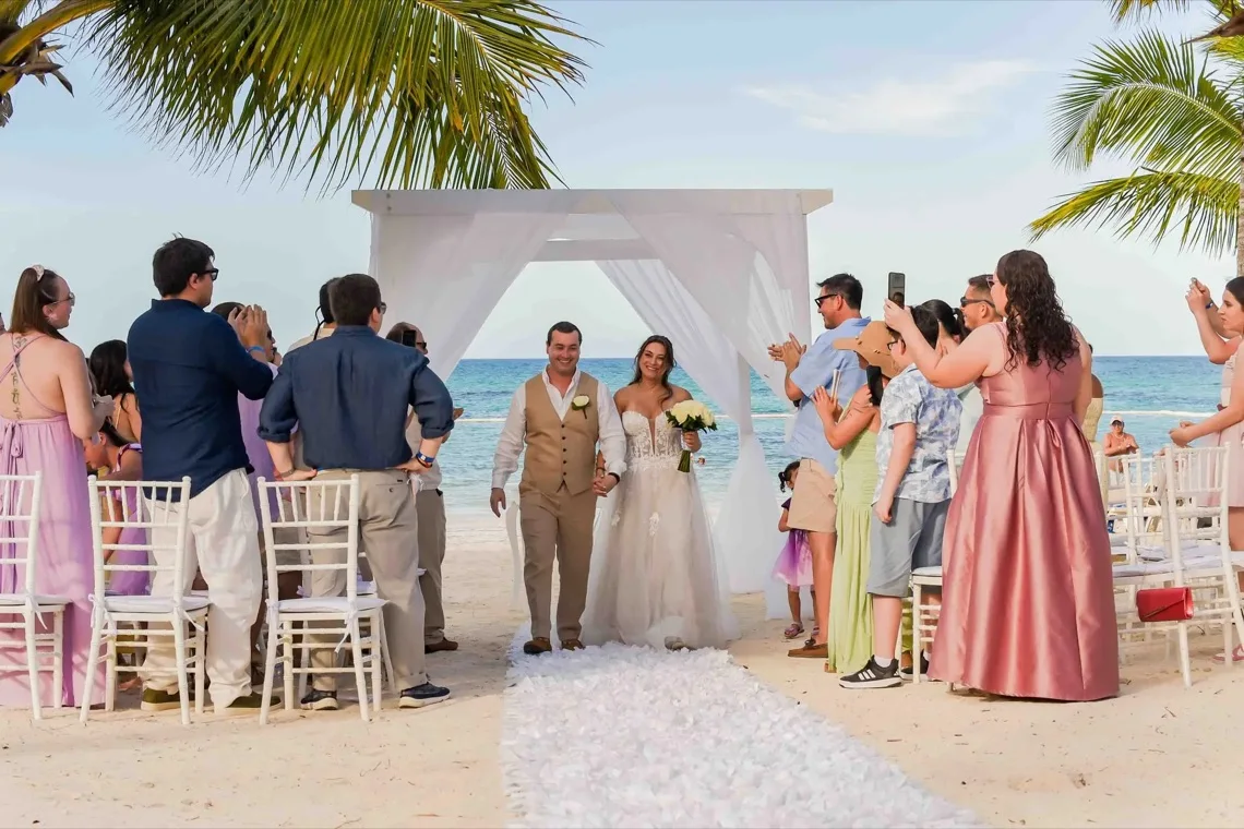 Novios caminando por el pasillo de una ceremonia en la playa rodeados de invitados