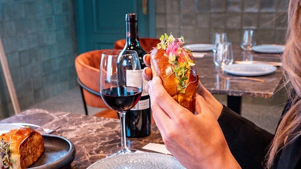 a person holding a sandwich with a bottle of wine in the background