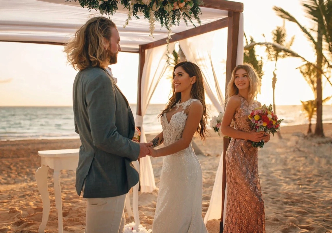 Novios y dama de honor bajo una pérgola en la playa al atardecer