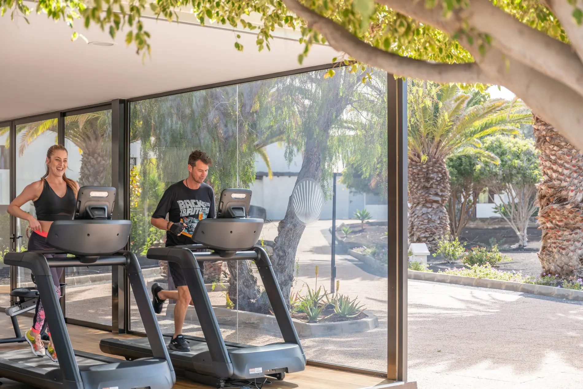 Casal a correr em passadeiras num ginásio envidraçado com vista para o jardim