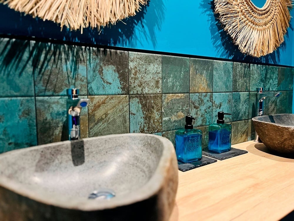 Baño de hotel moderno con lavabos de piedra, azulejos turquesa y decoración tropical.