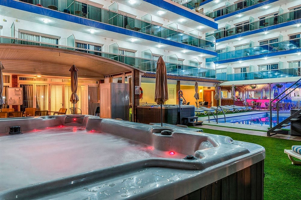 Relajante patio de hotel de noche con jacuzzi iluminado, piscina, bar y balcones.