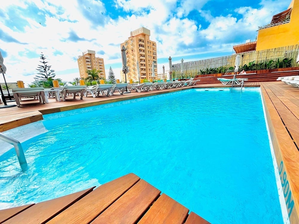 Piscina exterior de hotel soleada con terraza de madera, tumbonas y edificios de fondo.