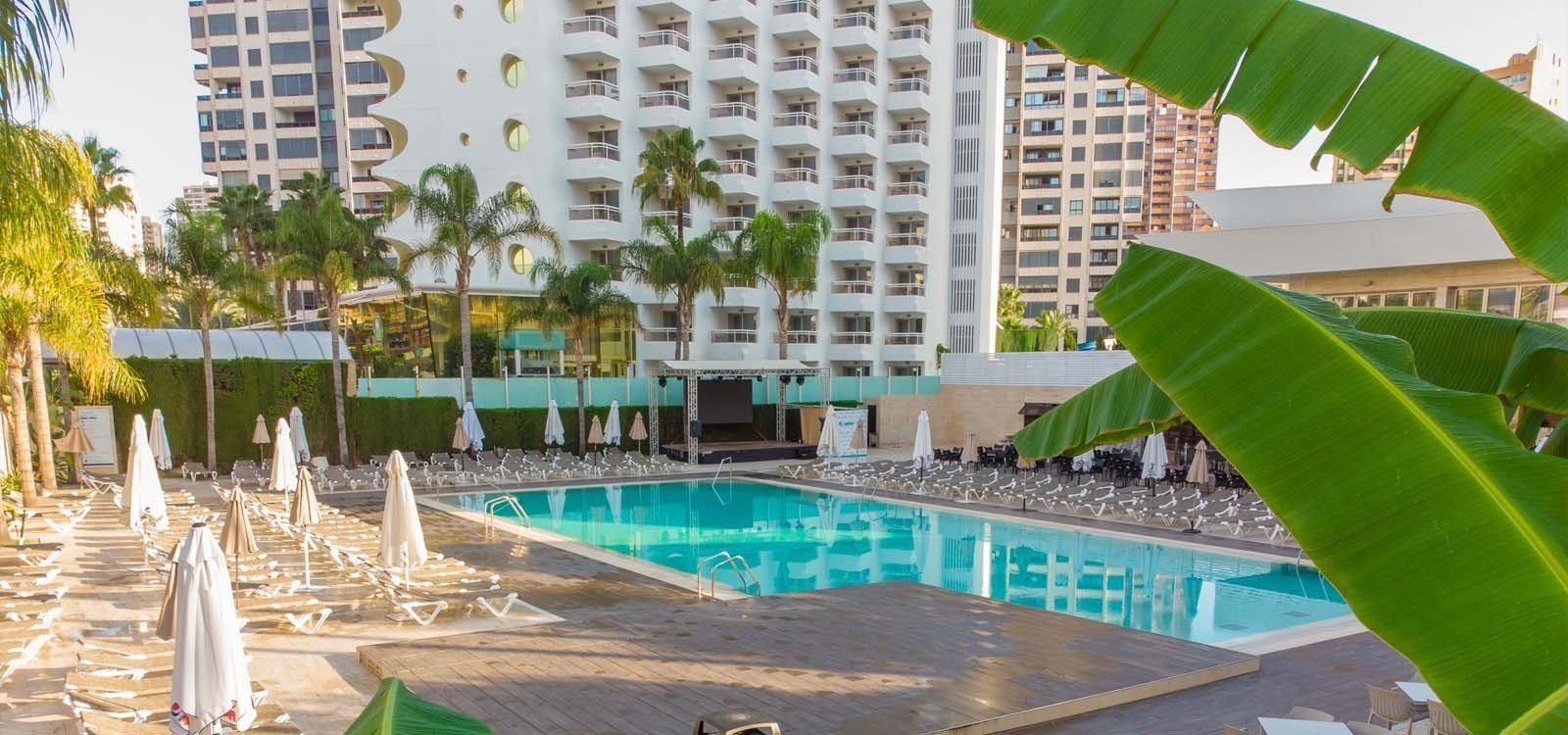 una piscina rodeada de sillas y sombrillas con un edificio en el fondo