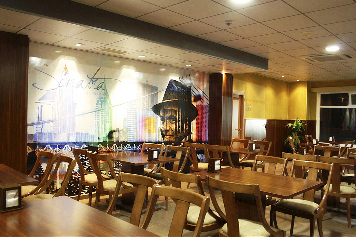 Interior de restaurante: mesas de madera, sillas y un mural de Frank Sinatra en un hotel. Hotel Acosta Ciudad de la Música