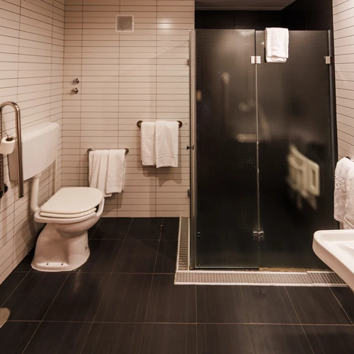 Modern hotel bathroom with white tiled walls, toilet, shower, and sink. Features accessible grab bar. Hotel Acosta Ciudad de la Música.