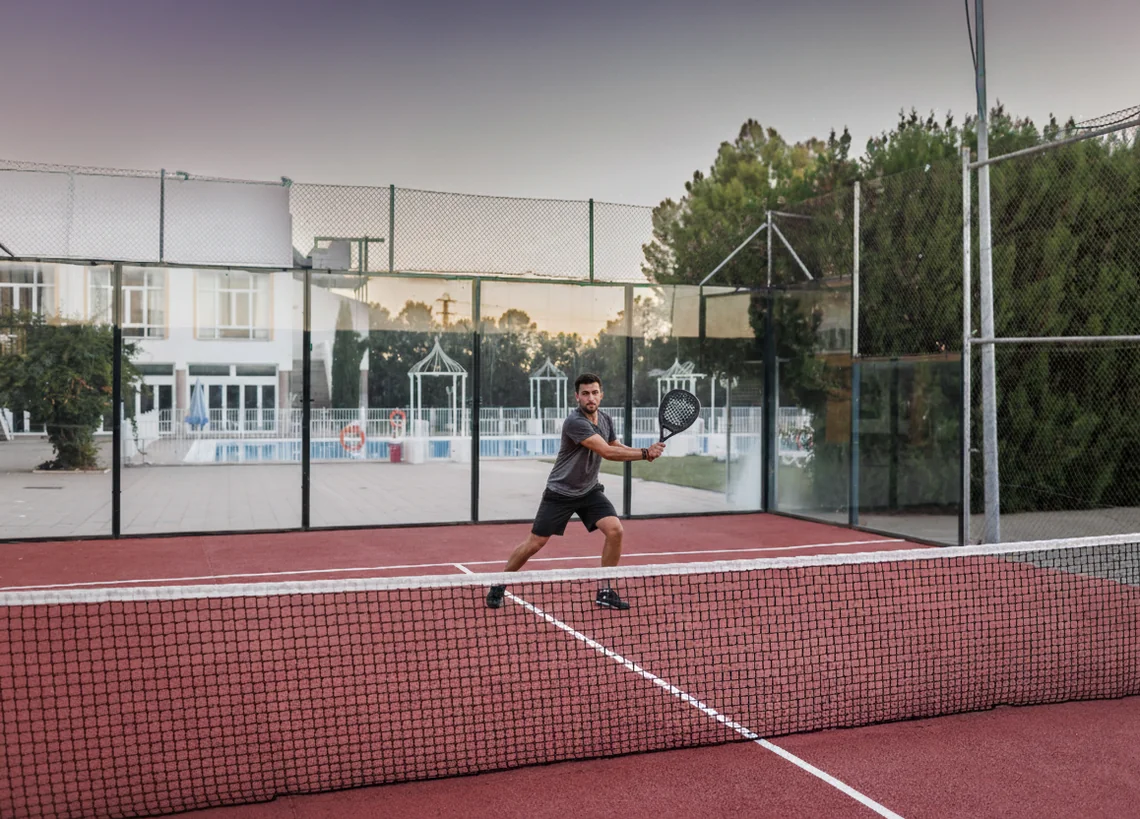 Man playing padel Hotel Acosta Ciudad de la Música on an outdoor court with a swimming pool and building in the background.