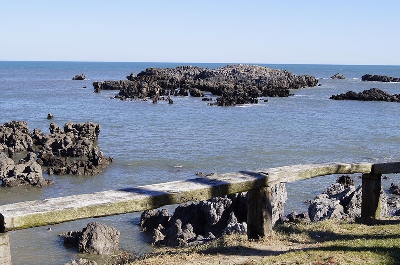 un banco de madera junto a un océano con rocas en el primer plano