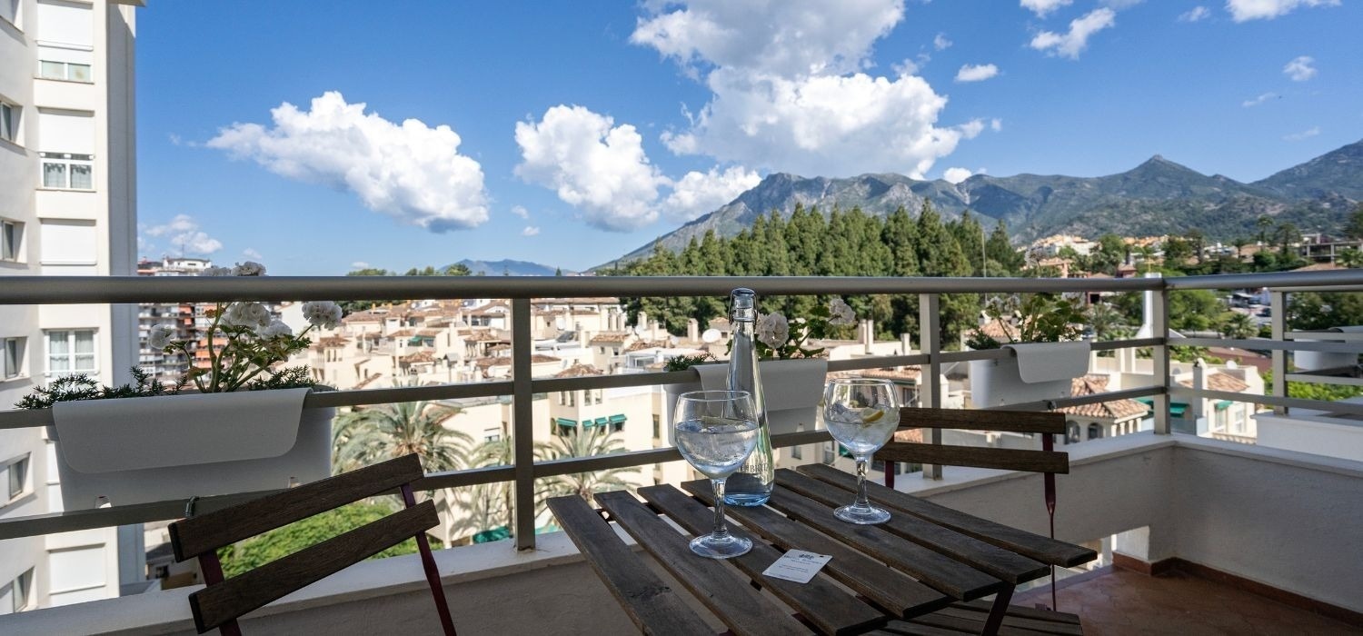 Un balcón soleado con una mesa y bebidas ofrece vistas panorámicas de montañas, árboles y tejados de un pueblo bajo un cielo azul con nubes.