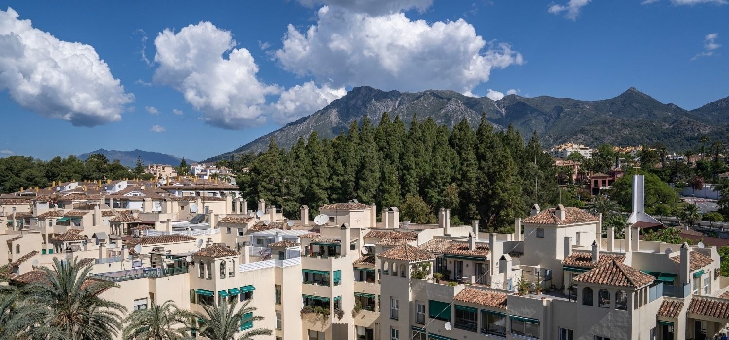 Un paisaje urbano de casas de tonos claros con tejados de terracota y abundante vegetación, enmarcado por montañas y un cielo azul con nubes.