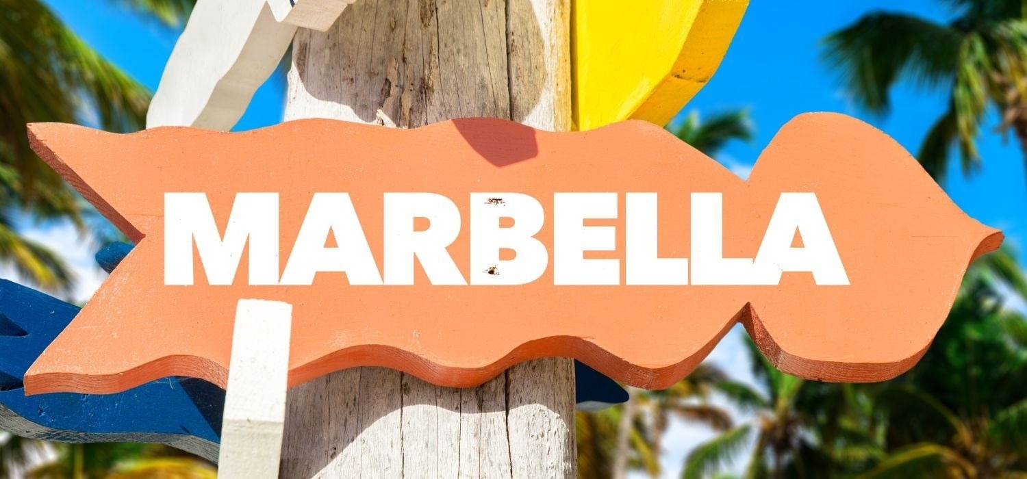 Un cartel direccional de madera, pintado de color naranja y con la palabra "MARBELLA" en letras blancas, se muestra contra un cielo azul y palmeras tropicales desenfocadas.