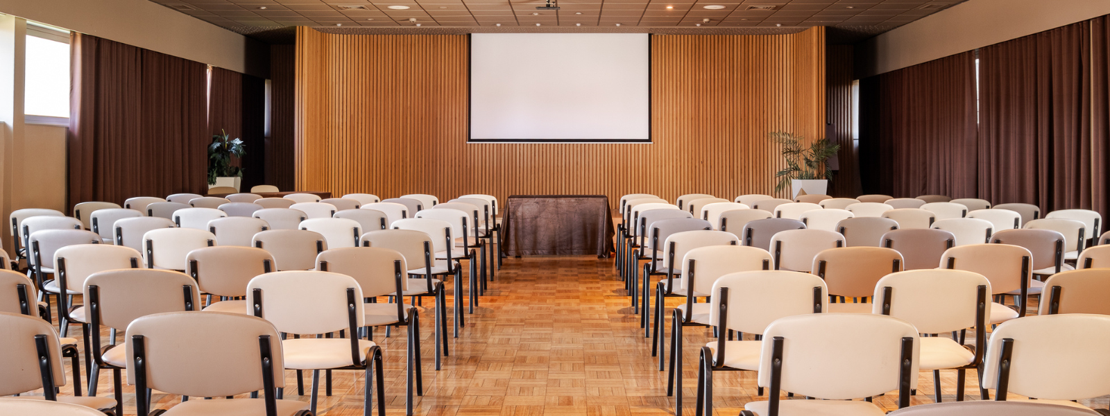 Une salle de conférence vide est équipée de rangées de chaises en bois clair, d'un écran de projection et d'un paperboard, avec des murs en lambris de bois et un sol carrelé sombre.