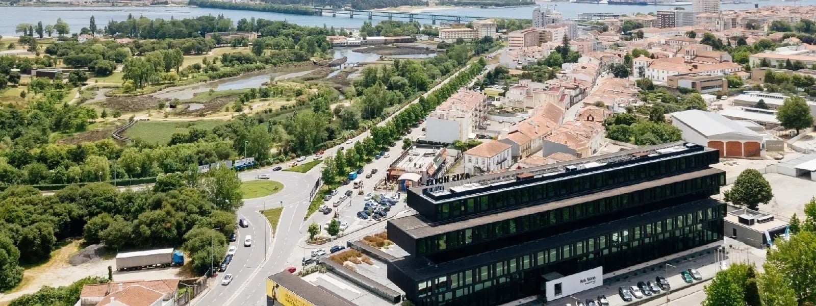 A imagem apresenta uma vista aérea de uma cidade com um edifício moderno de tons escuros em primeiro plano, rodeado por áreas verdes, ruas com tráfego e um rio com uma ponte ao fundo.