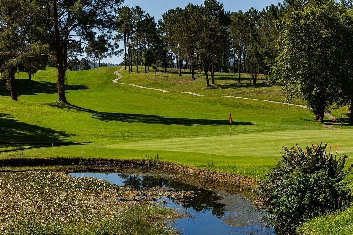 Un terrain de golf verdoyant est visible avec un étang rempli de nénuphars au premier plan, un drapeau rouge sur le green et des arbres bordant le parcours sous un ciel clair.