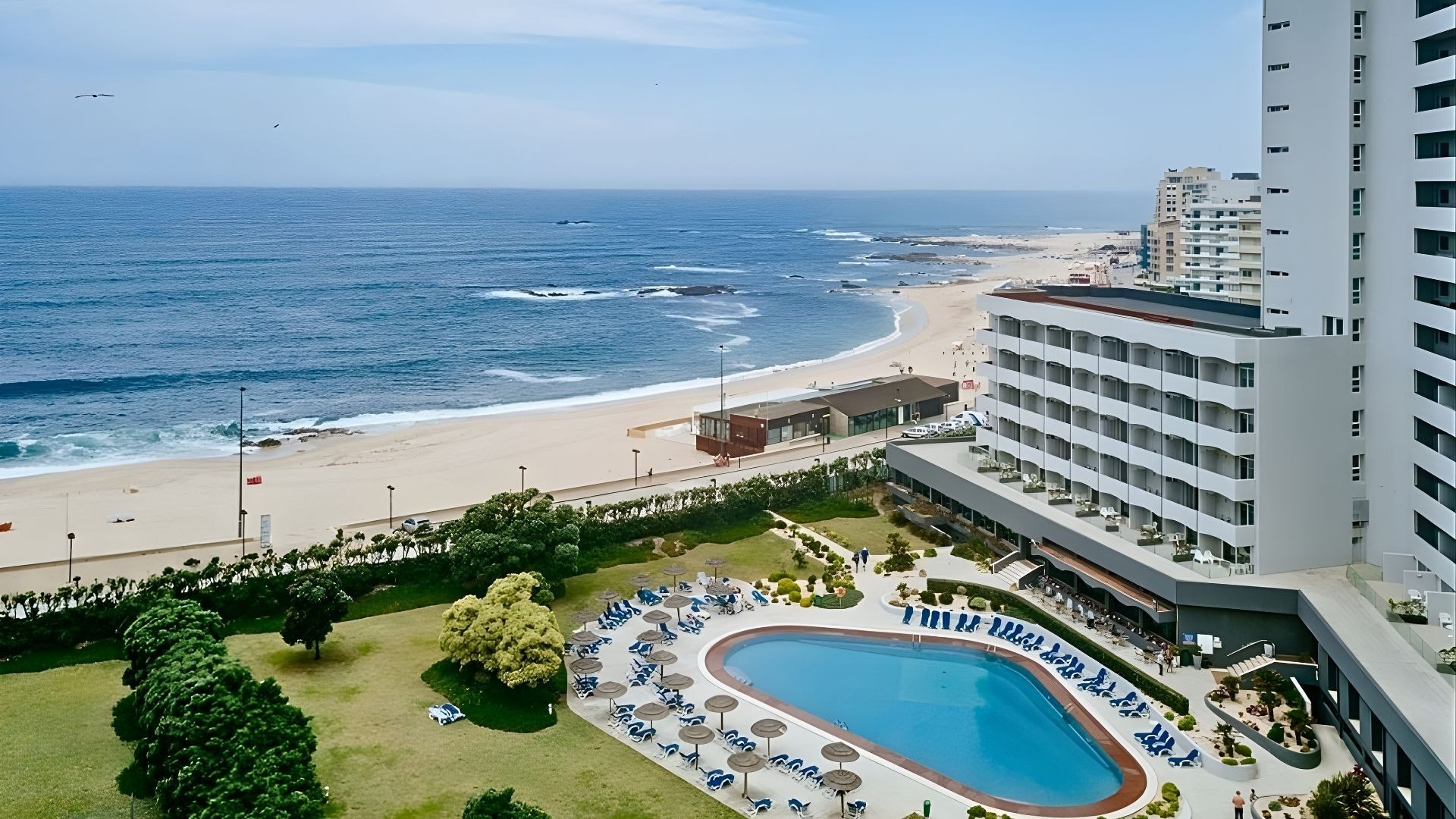 Vue aérienne d'un complexe hôtelier moderne avec piscine et pelouses, situé en bord de mer le long d'une plage de sable sous un ciel légèrement nuageux.