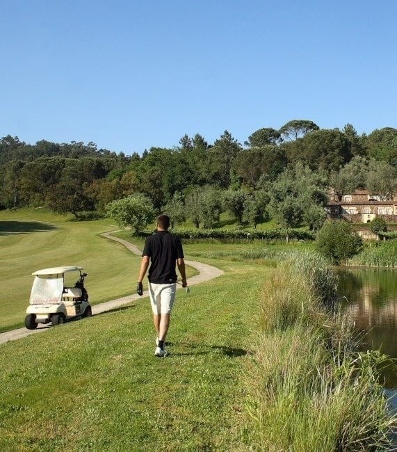 Un golfeur marche sur un parcours verdoyant, accompagné d'une voiturette, le long d'un lac et de forêts lointaines, sous un ciel bleu.