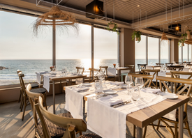 Une salle de restaurant lumineuse et élégante avec de nombreuses tables dressées est visible à travers de grandes baies vitrées donnant sur une végétation luxuriante et ensoleillée.