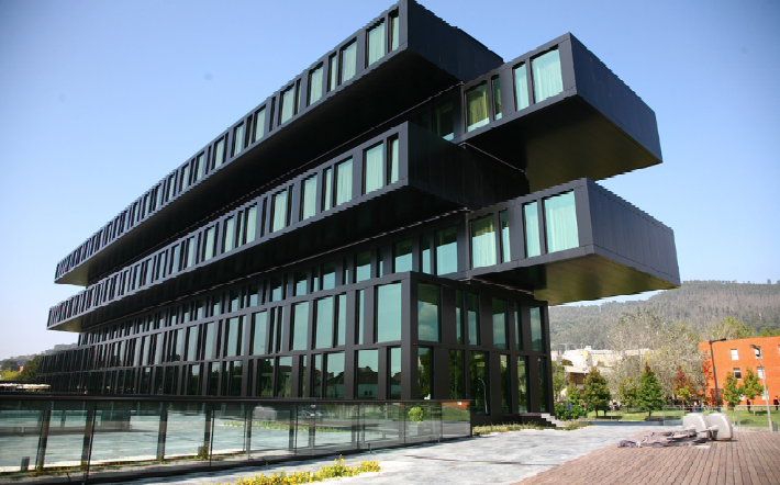 Um edifício moderno e escuro com design geométrico, apresentando blocos sobrepostos e em balanço com muitas janelas, destaca-se sob um céu azul claro.