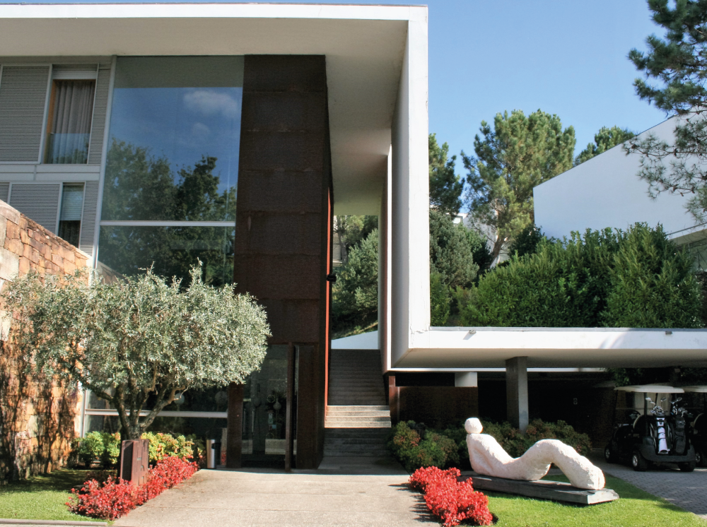 Uma casa moderna de arquitetura marcante com janelas espelhadas, elementos brancos e marrons, rodeada por vegetação e adornada por uma escultura reclinada no jardim.