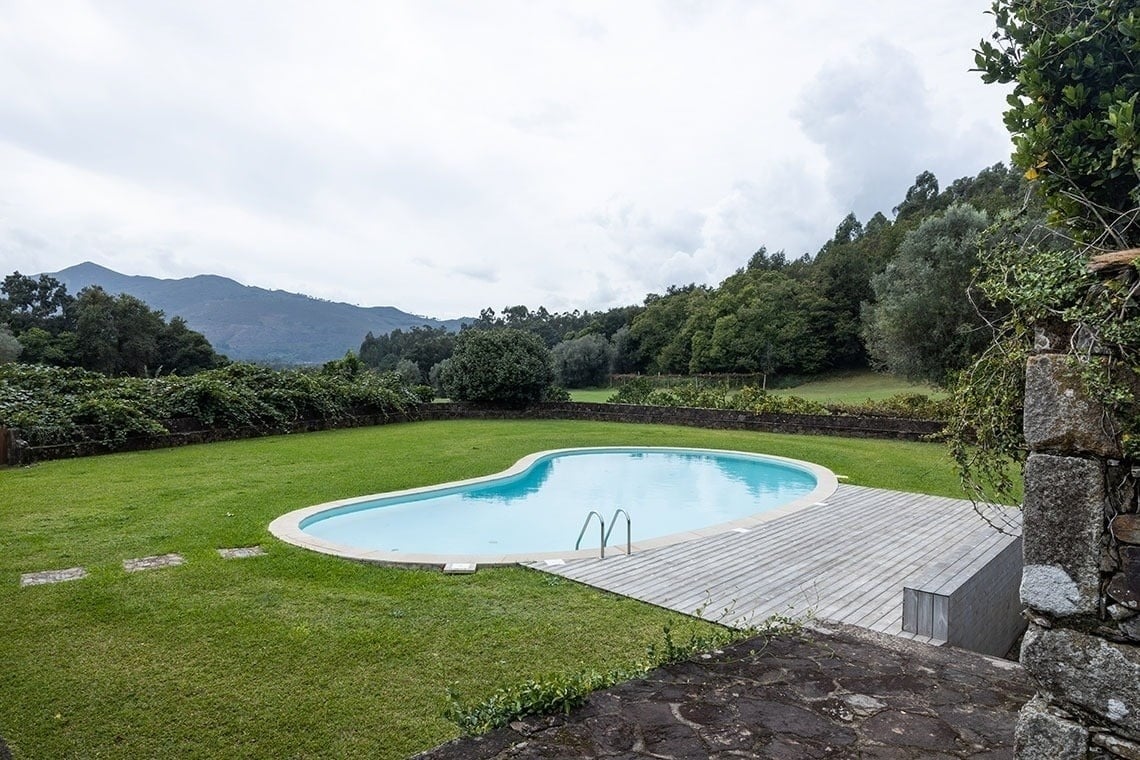 Uma piscina de formato orgânico com deck de madeira e gramado verde, com montanhas e floresta ao fundo sob um céu nublado.
