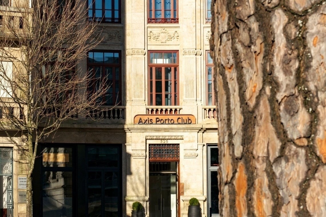 A fachada de um edifício com o letreiro "Axis Porto Club" é visível, parcialmente obscurecida por uma árvore sem folhas à esquerda e um tronco de árvore à direita.