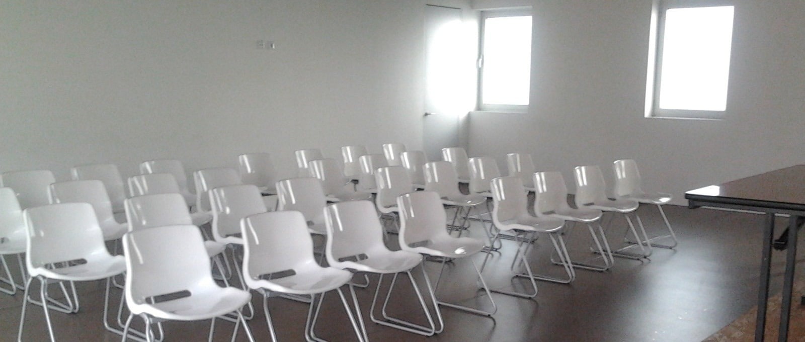 Une salle de conférence vide est équipée de rangées de chaises en bois clair, d'un écran de projection et d'un paperboard, avec des murs en lambris de bois et un sol carrelé sombre.