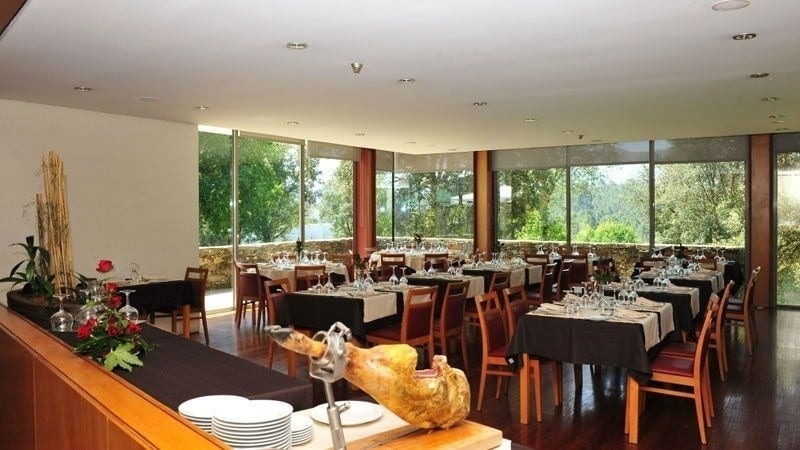 Une salle de restaurant lumineuse et élégante avec de nombreuses tables dressées est visible à travers de grandes baies vitrées donnant sur une végétation luxuriante et ensoleillée.