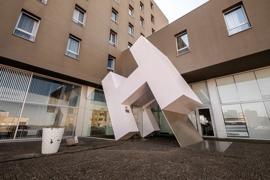 Une structure architecturale blanche et angulaire, ressemblant à un "H" stylisé, sert d'entrée à un bâtiment moderne de couleur taupe avec de nombreuses fenêtres.